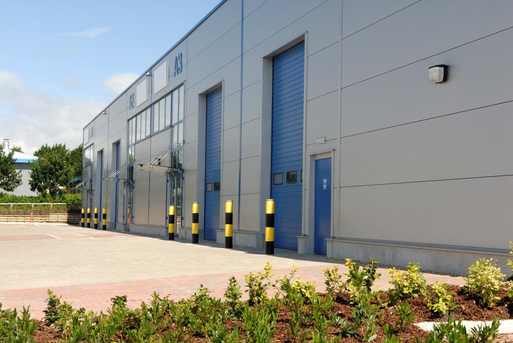 Modern industrial warehouse building with gray metal siding, tall blue roll-up loading doors, and yellow-and-black safety bollards along a paved driveway, with landscaped shrubs in the foreground