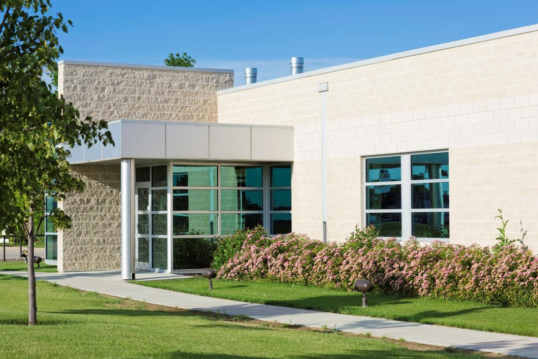Modern single-story commercial building with large windows, light brick exterior, landscaped shrubs, and a sidewalk leading to the entrance on a sunny day.