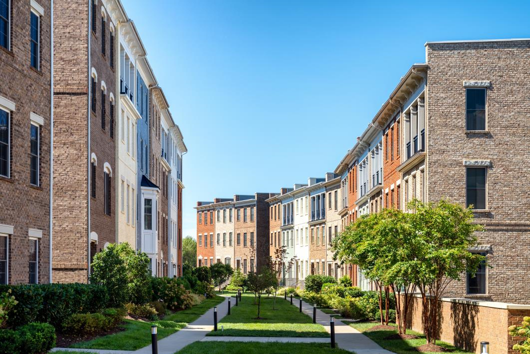 Row of modern brick townhouses with landscaped green space and walking paths between buildings under a clear blue sky.
