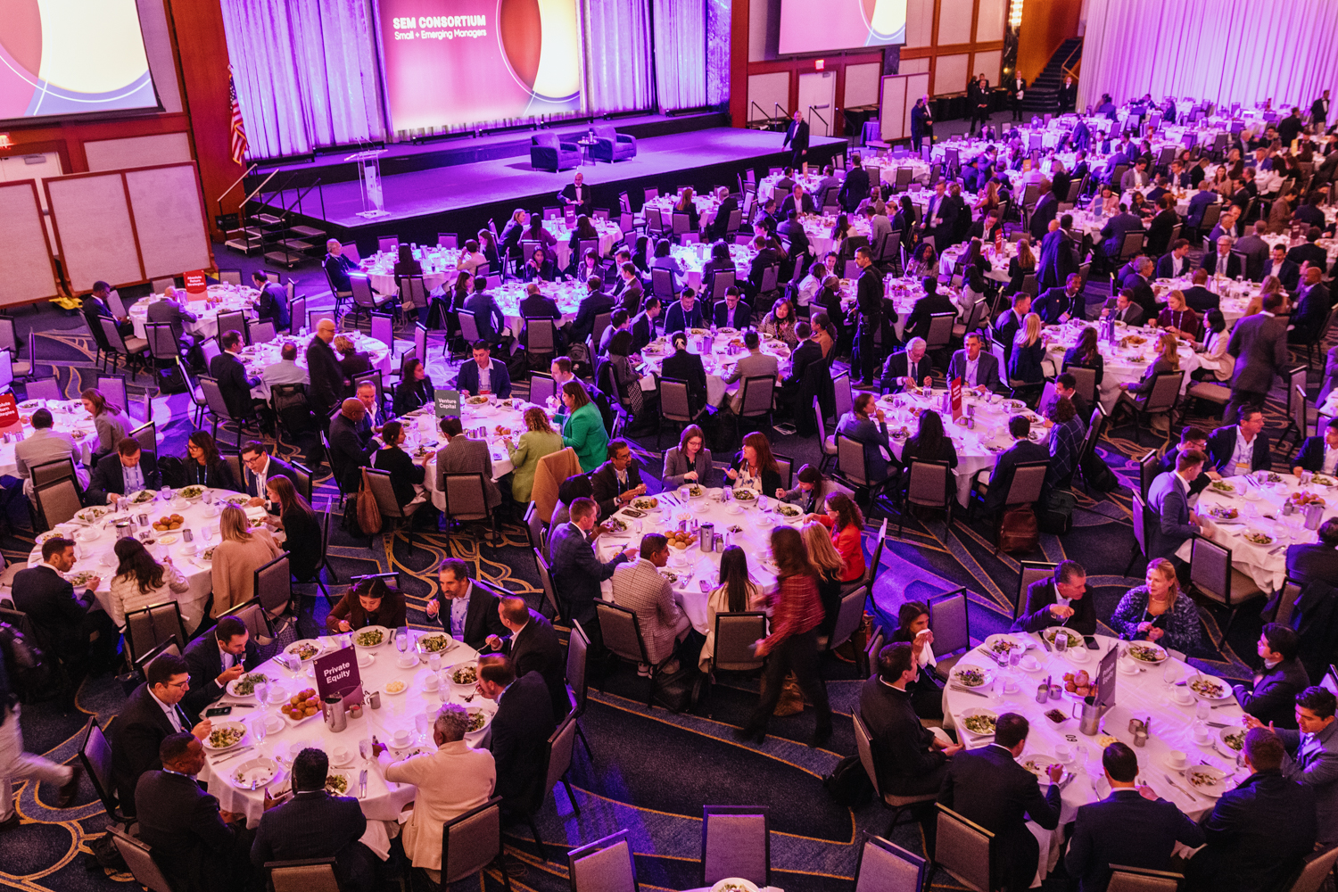 Large conference banquet with attendees seated at round tables in a ballroom, facing a stage with presentation screens and purple lighting.