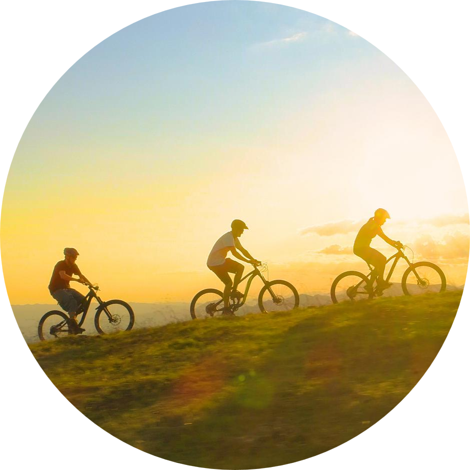 Three cyclists riding mountain bikes along a grassy ridge at sunset, silhouetted against a golden sky.
