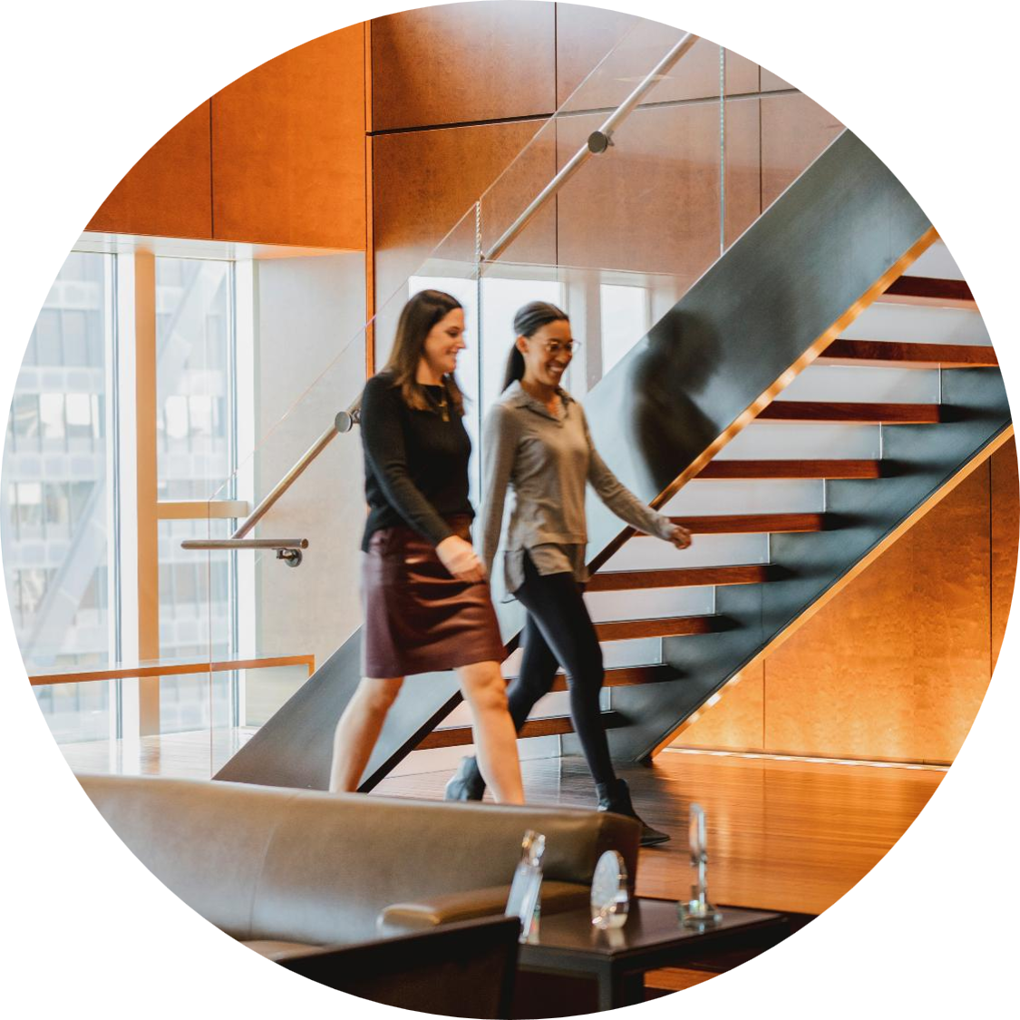 Two colleagues walking and smiling inside a modern office lobby, passing in front of a glass-and-metal staircase with wood-paneled walls and large windows.