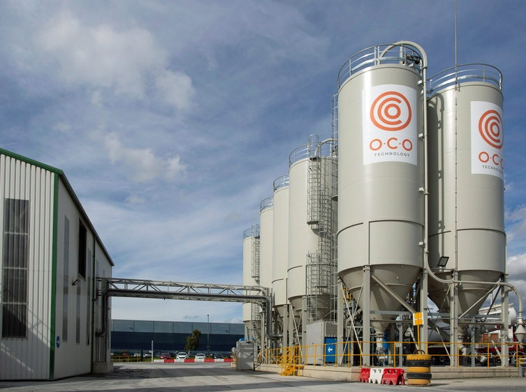 Industrial carbon capture facility with multiple tall white storage silos labeled “O.C.O Technology,” connected by metal piping and walkways under a partly cloudy sky.