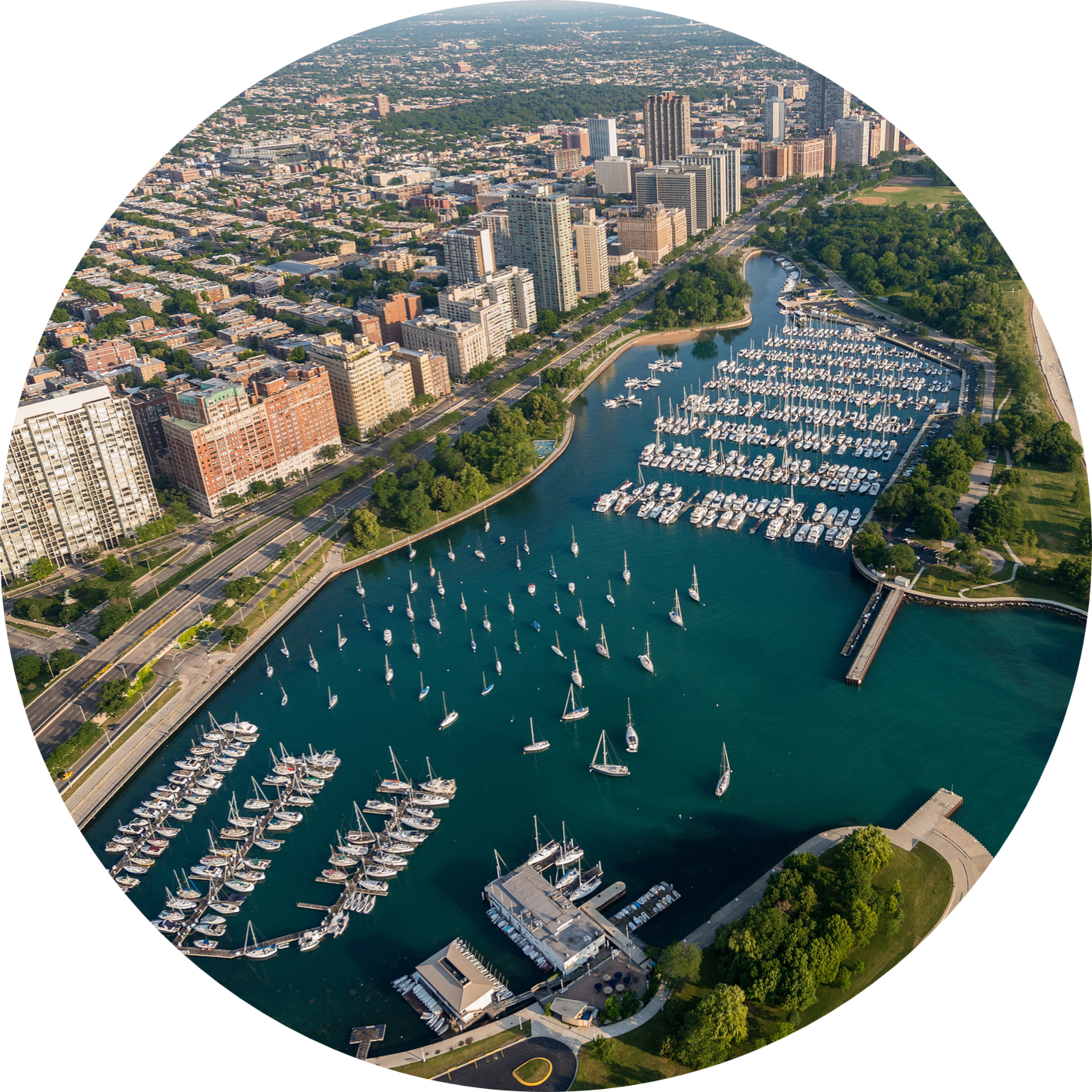 Aerial view of a city waterfront with marinas filled with sailboats, high-rise buildings, and a tree-lined shoreline.