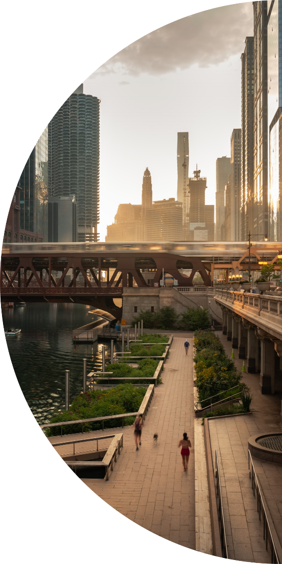 Riverfront cityscape at sunset with a pedestrian walkway, bridge, and modern high-rise buildings in the background.